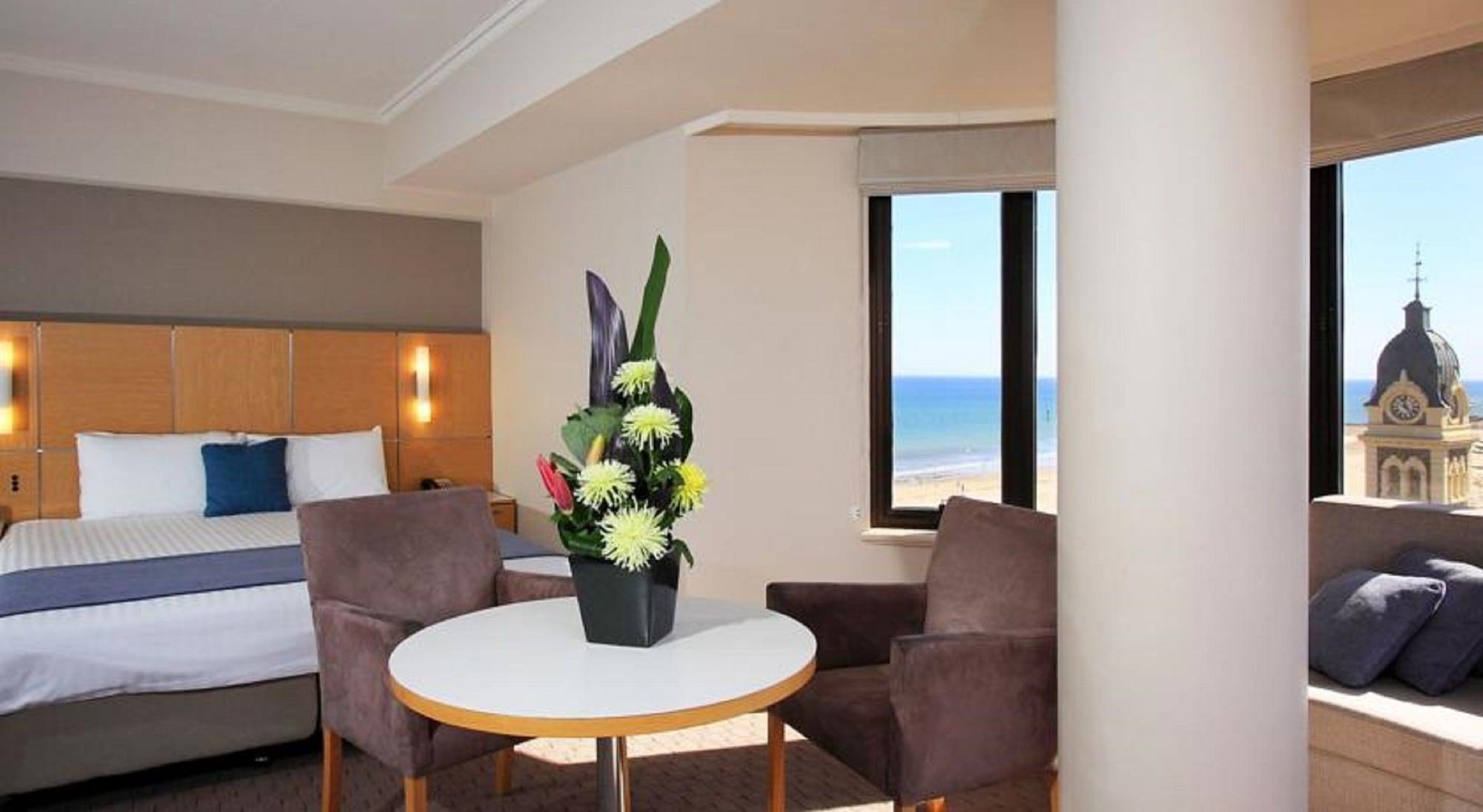 Hotel Stamford Grand Adelaide à Glenelg Extérieur photo The photo shows a cozy hotel room with a comfortable seating arrangement. There are two brown chairs positioned around a round table, which features a decorative flower arrangement in a black vase. The background includes a window that offers a view 