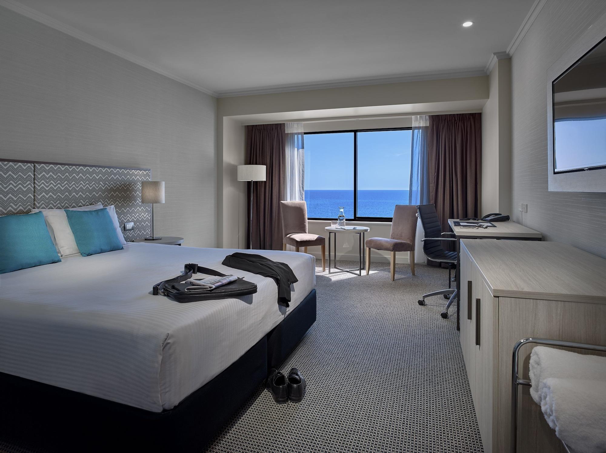 Hotel Stamford Grand Adelaide à Glenelg Extérieur photo The photo shows a modern hotel room featuring a queen-sized bed with white linens and two aqua blue decorative pillows. There are bedside lamps and a large window with a view of the ocean. The room also has a small seating area with two chairs and a 