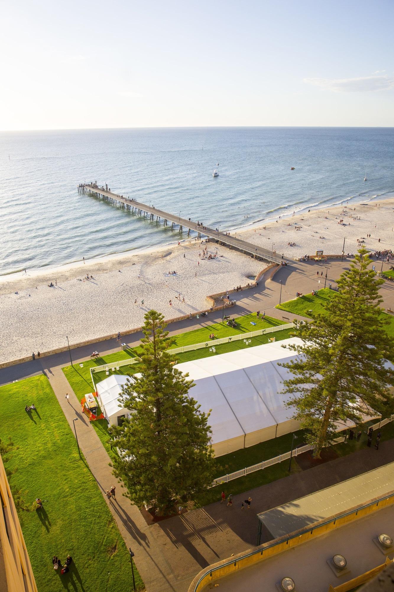 Hotel Stamford Grand Adelaide à Glenelg Extérieur photo The photo shows a scenic view of a beach with a long pier extending into the water. The beach is sandy, and there are people enjoying the sun and walking along the shore. In the foreground, there is a large white tent set up on a grassy area, likely 