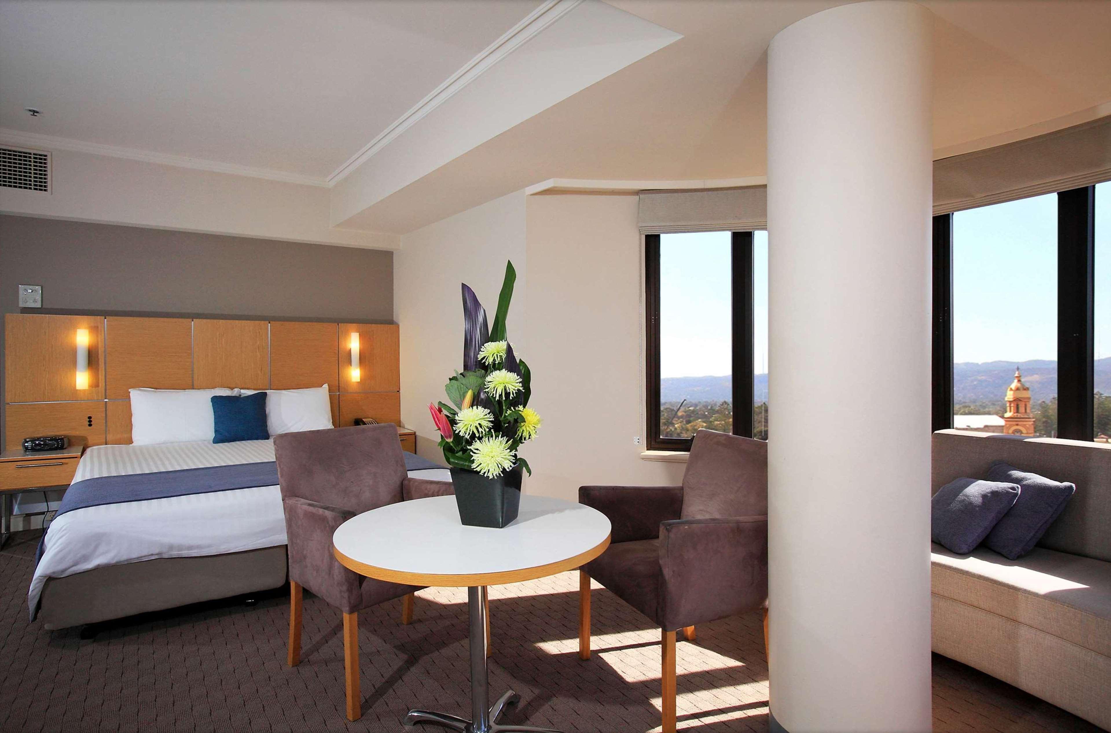 Hotel Stamford Grand Adelaide à Glenelg Extérieur photo The photo shows a hotel room with a modern design. On one side, there is a plush bed with white and blue bedding. In the center of the room, there's a small round table with two gray upholstered chairs around it. A vase of flowers sits on the table, 