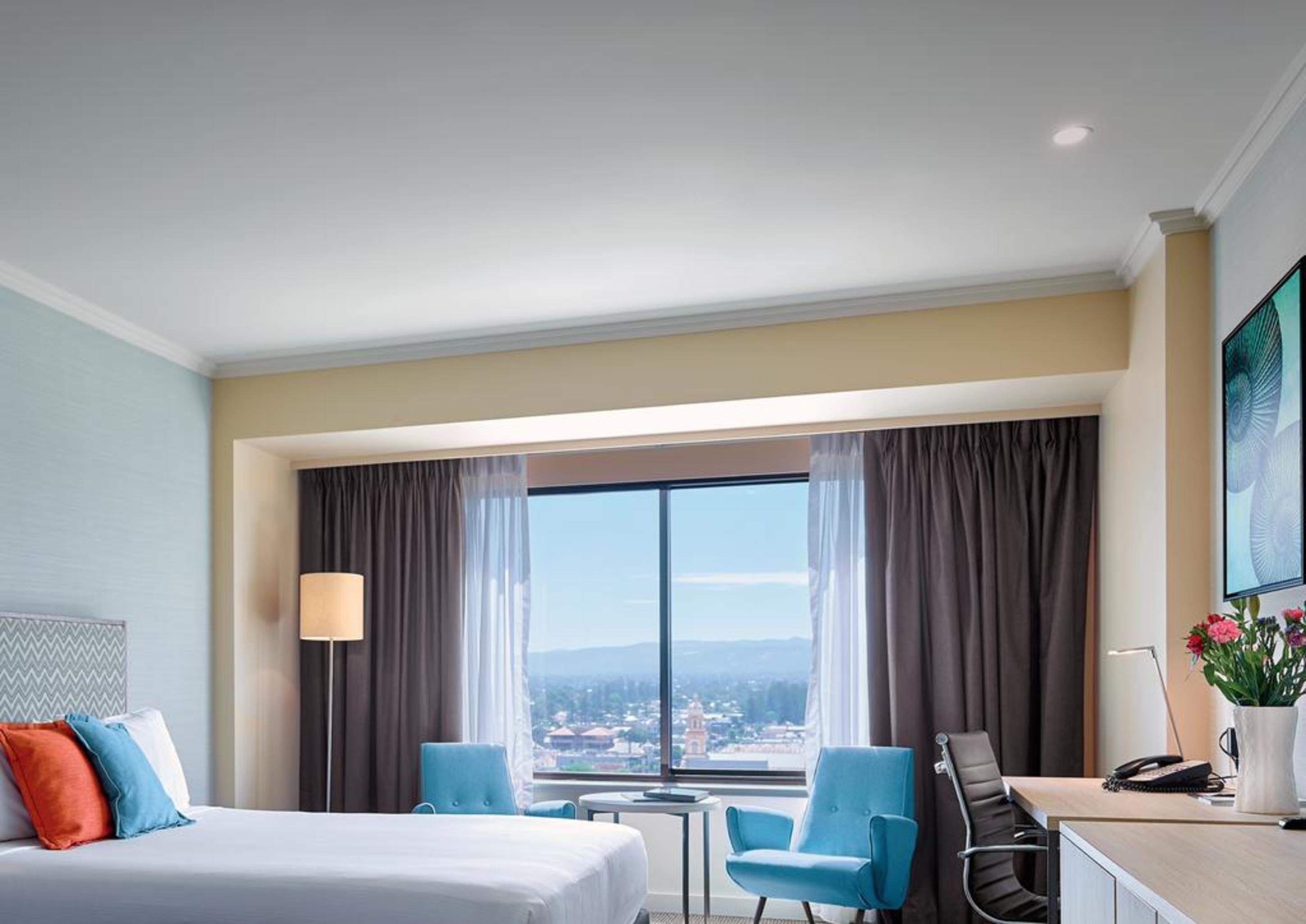 Hotel Stamford Grand Adelaide à Glenelg Extérieur photo The photo shows a modern hotel room featuring a queen-sized bed with white linens and colorful decorative pillows. There are two blue chairs beside a small table positioned near a large window that provides a view of the outside, with sheer and dark 