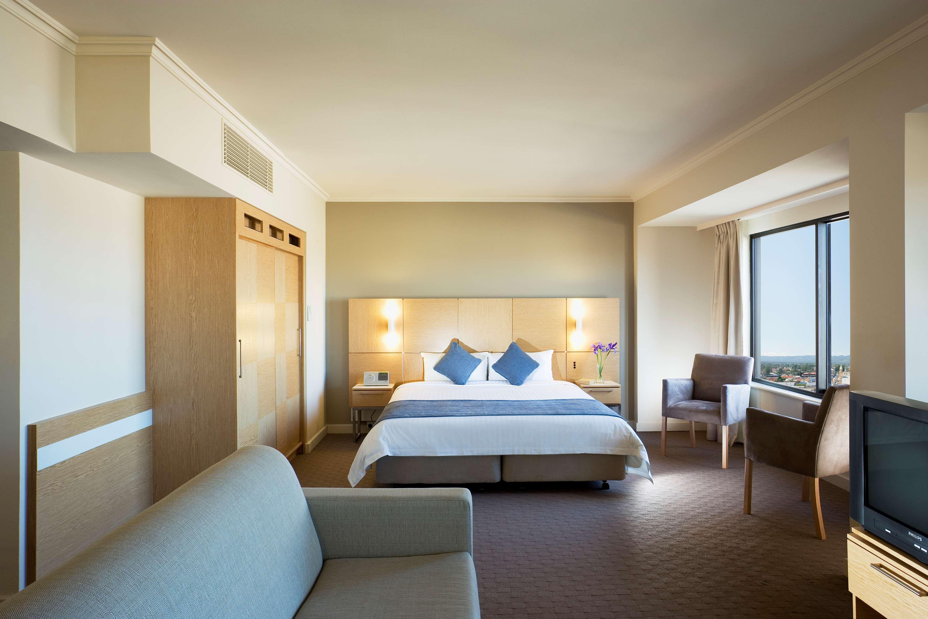 Hotel Stamford Grand Adelaide à Glenelg Chambre photo The photo shows a modern hotel room featuring a large bed with two blue decorative pillows. There are bedside lamps on both sides of the headboard. To the left, there's a wooden wardrobe and a small table with a vase and a few amenities. On the right
