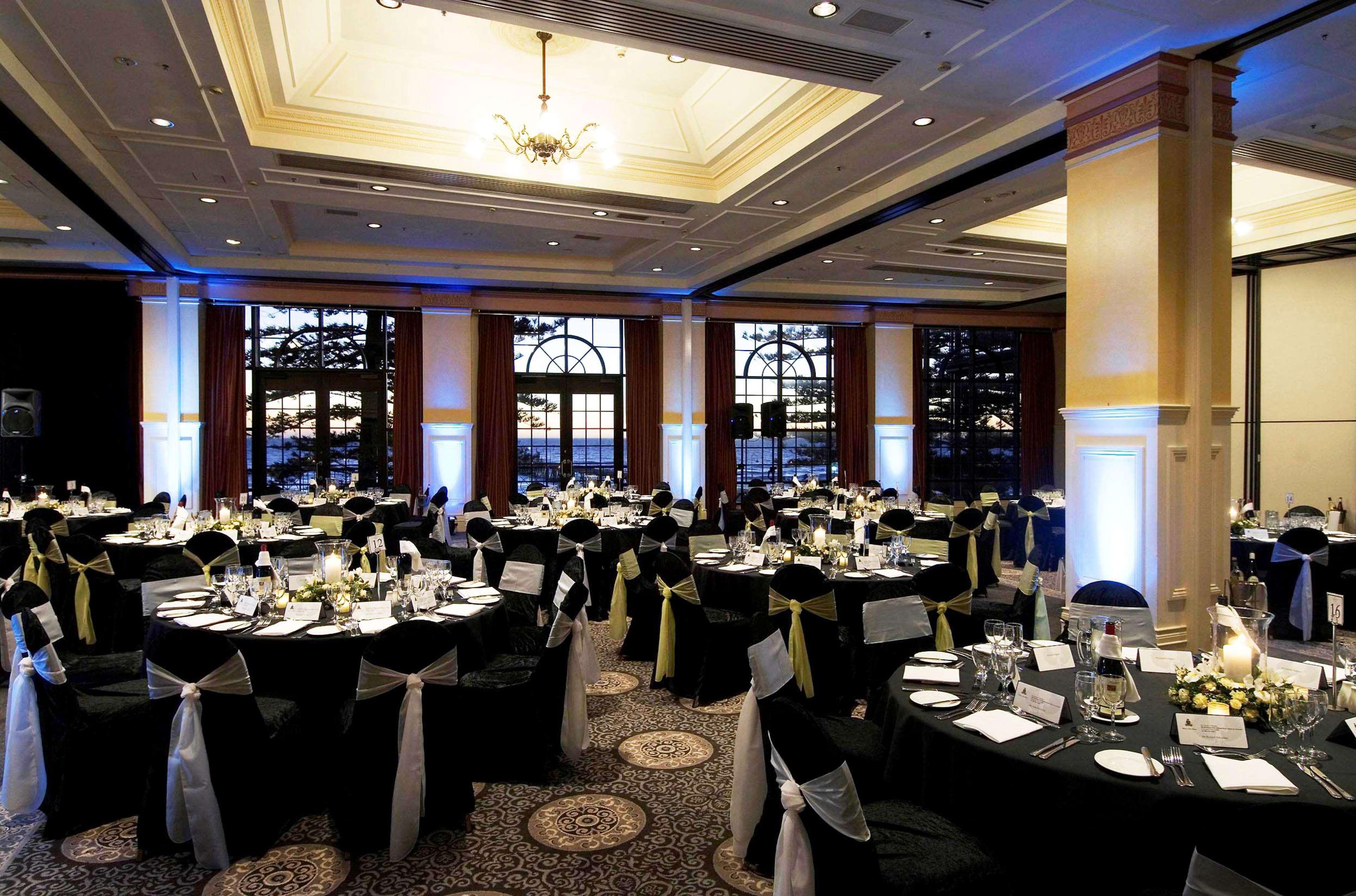 Hotel Stamford Grand Adelaide à Glenelg Restaurant photo The photo depicts an elegantly arranged banquet hall set for an event. The room features round tables covered with black tablecloths, each adorned with white tableware and floral centerpieces. Chairs are draped in white fabric with decorative sashes 