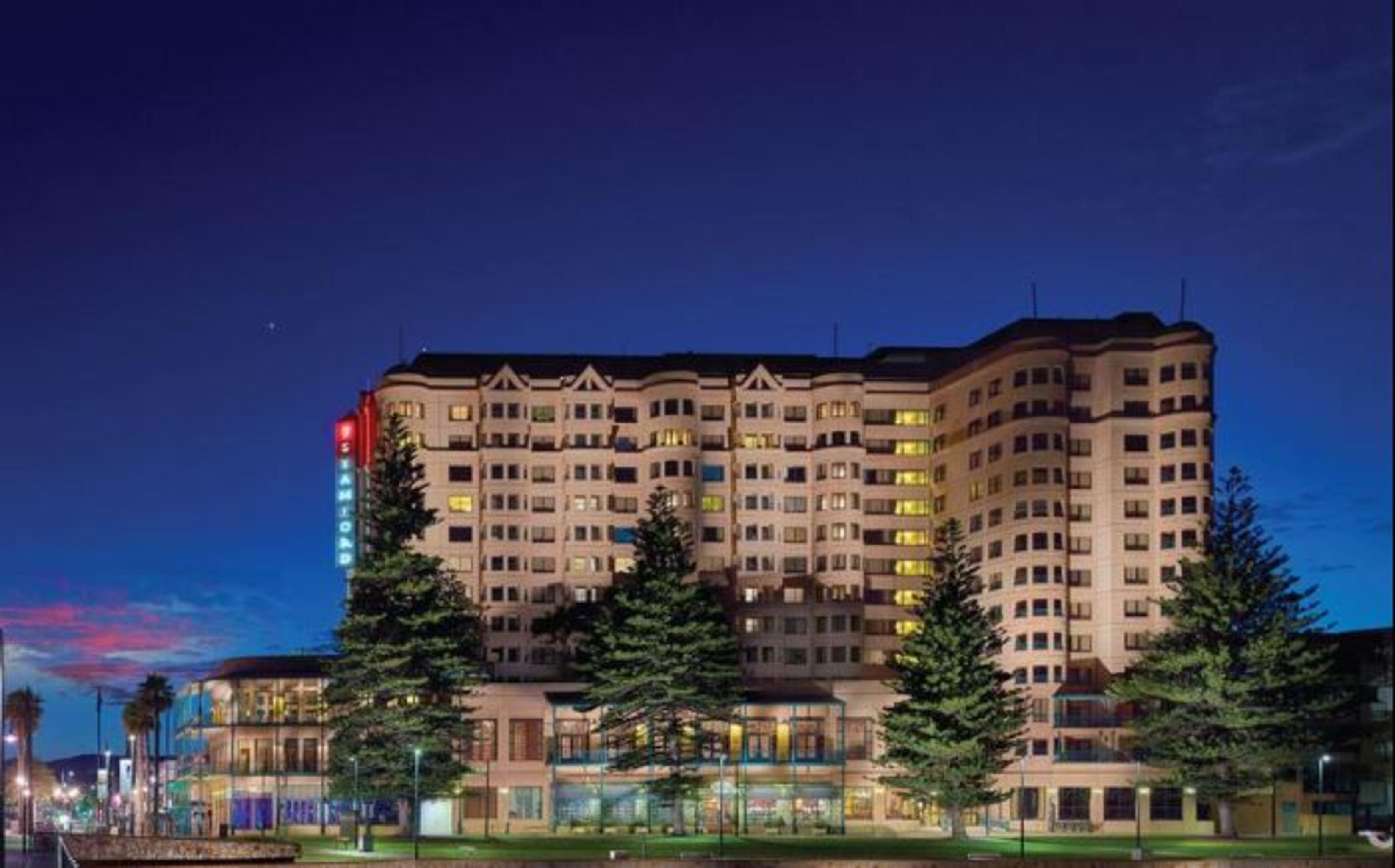 Hotel Stamford Grand Adelaide à Glenelg Extérieur photo The photo displays a multi-story hotel building at dusk, illuminated by various lights. The architecture features a mix of modern and traditional design elements, with numerous windows showcasing a warm glow. Surrounding the hotel are several tall tr
