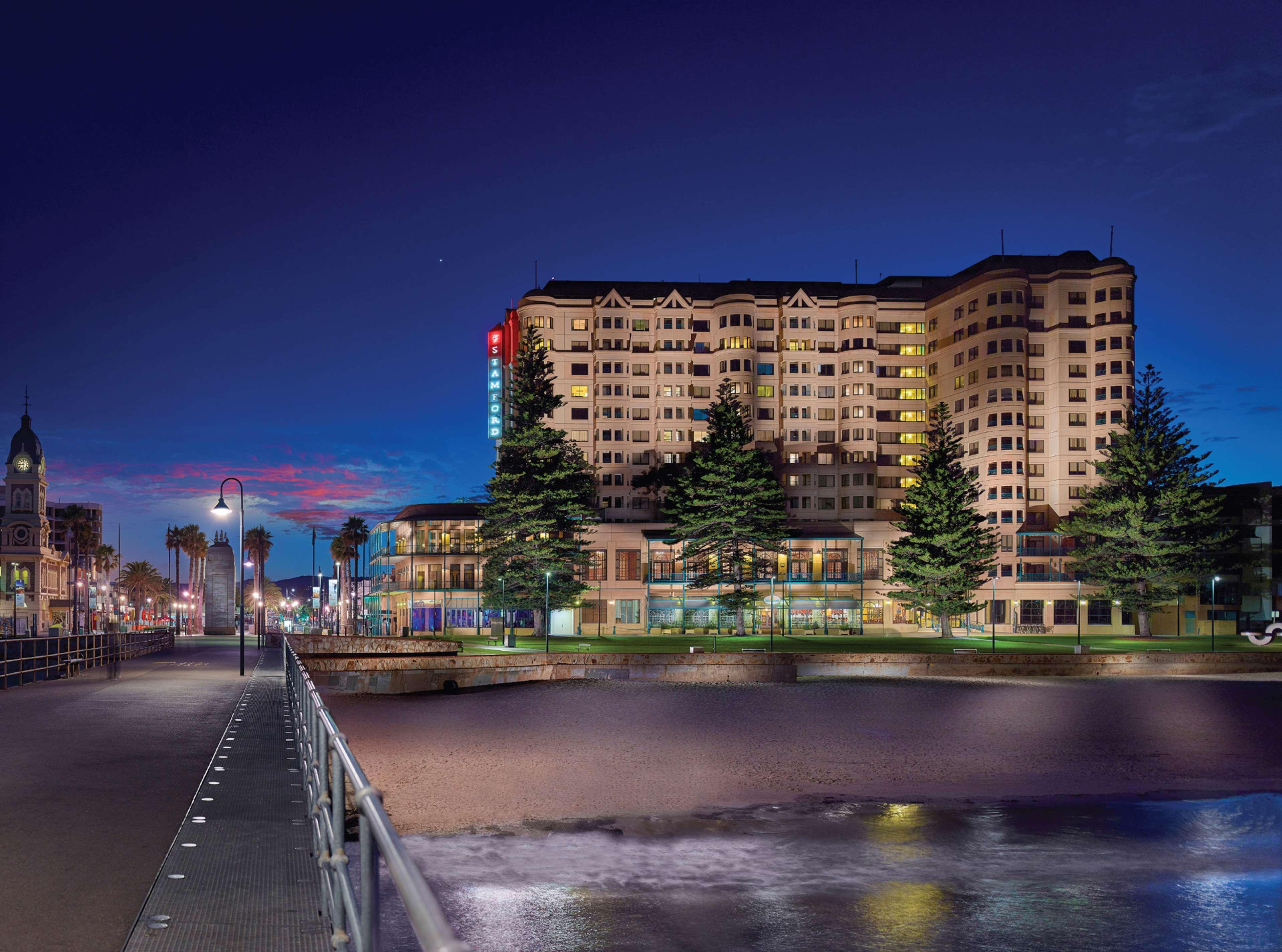 Hotel Stamford Grand Adelaide à Glenelg Extérieur photo The photo shows a waterfront scene at dusk, featuring a large building lit up against a darkening sky. The architecture appears modern and prominent, with several windows illuminated. In the foreground, there is a walkway or pier with fencing along t