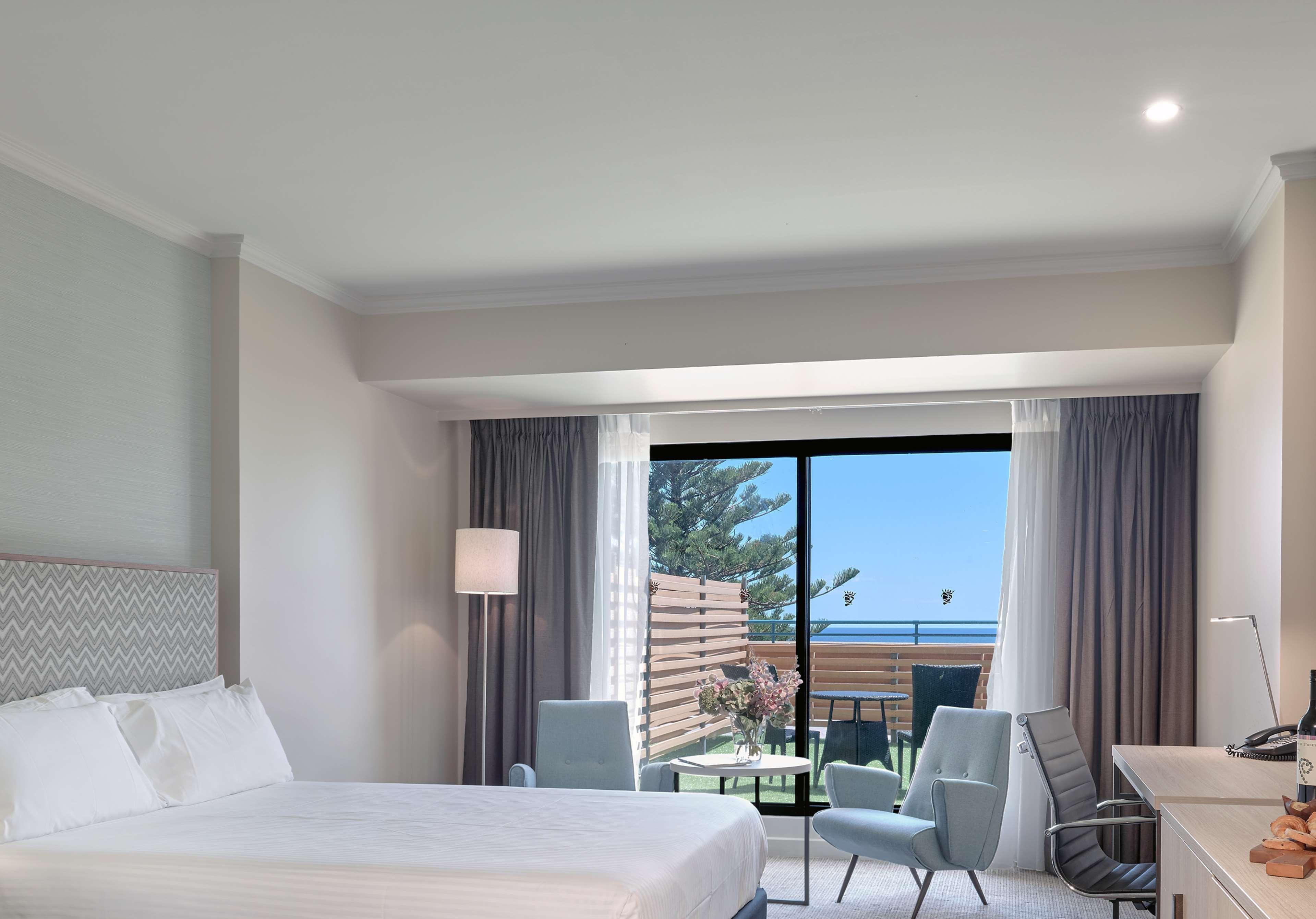 Hotel Stamford Grand Adelaide à Glenelg Extérieur photo The photo shows a modern hotel room featuring a large bed with white bedding. There are two armchairs positioned near a window that offers a view of an outdoor area, possibly a balcony or patio with greenery. The room has light-colored walls and curt