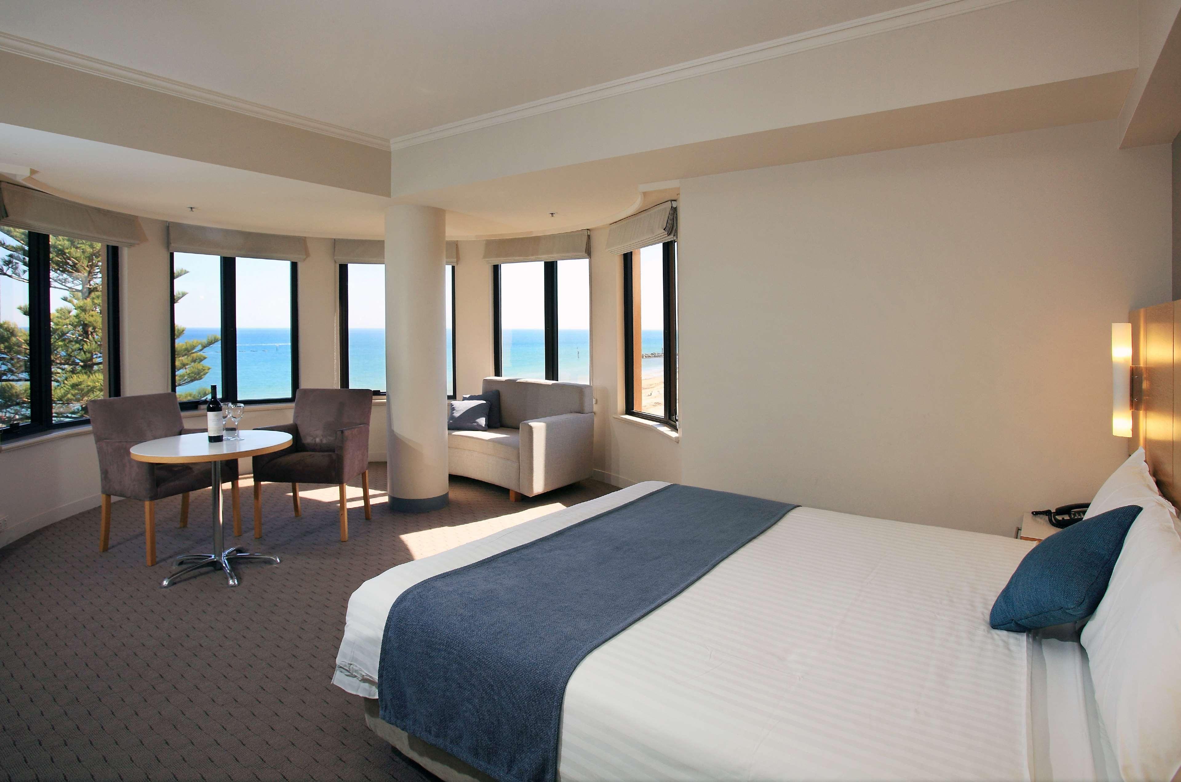 Hotel Stamford Grand Adelaide à Glenelg Extérieur photo The photo shows a well-lit hotel room featuring large windows that offer a view of the ocean. The room is arranged with a comfortable bed covered in white linens and a navy blue throw blanket. There is a small table and chairs set up beside the windo