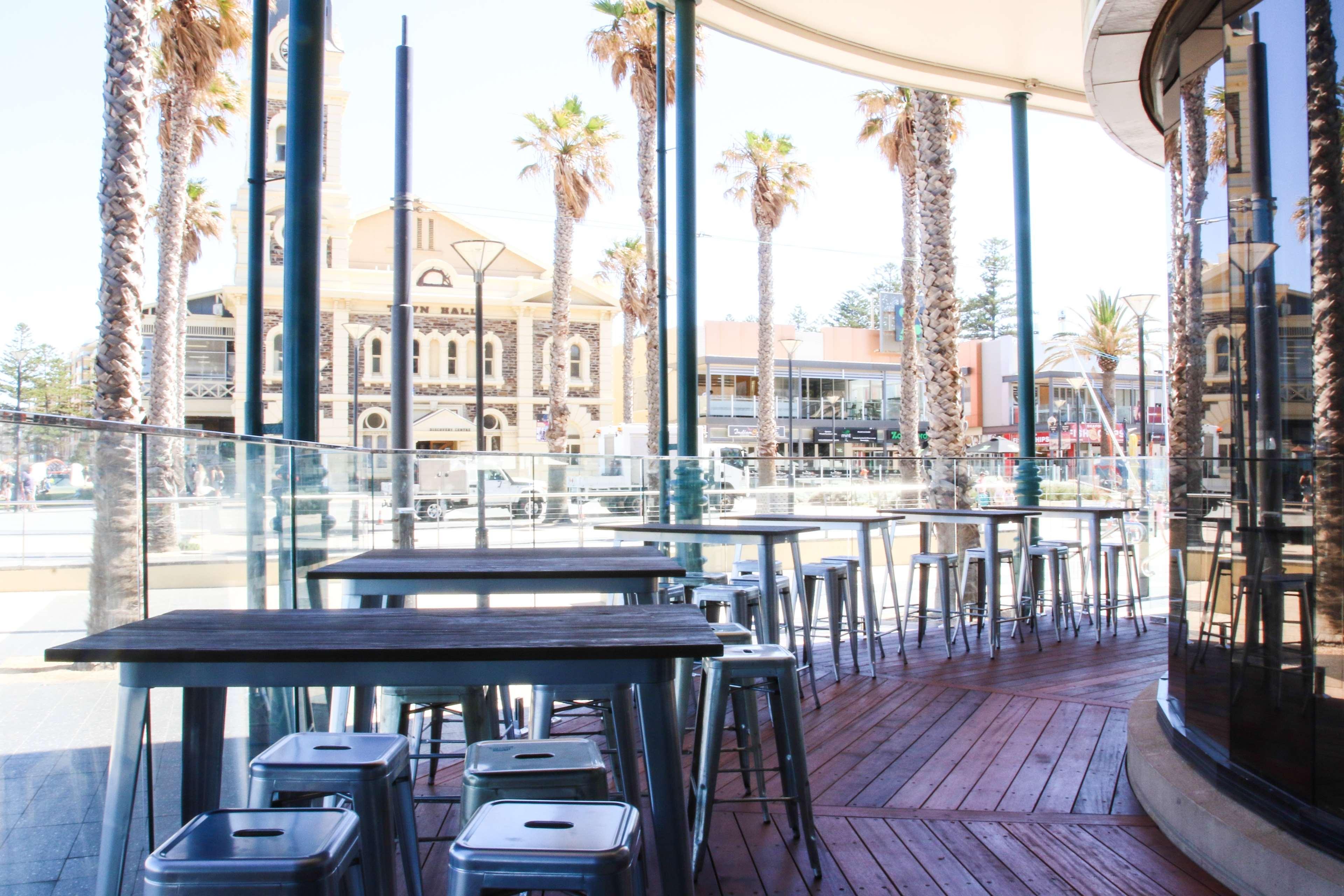 Hotel Stamford Grand Adelaide à Glenelg Extérieur photo The photo shows an outdoor seating area of a café or restaurant. It features metal tables and stools arranged on a wooden deck. The setting has large glass panels, providing an open and airy atmosphere. In the background, palm trees and a building wi
