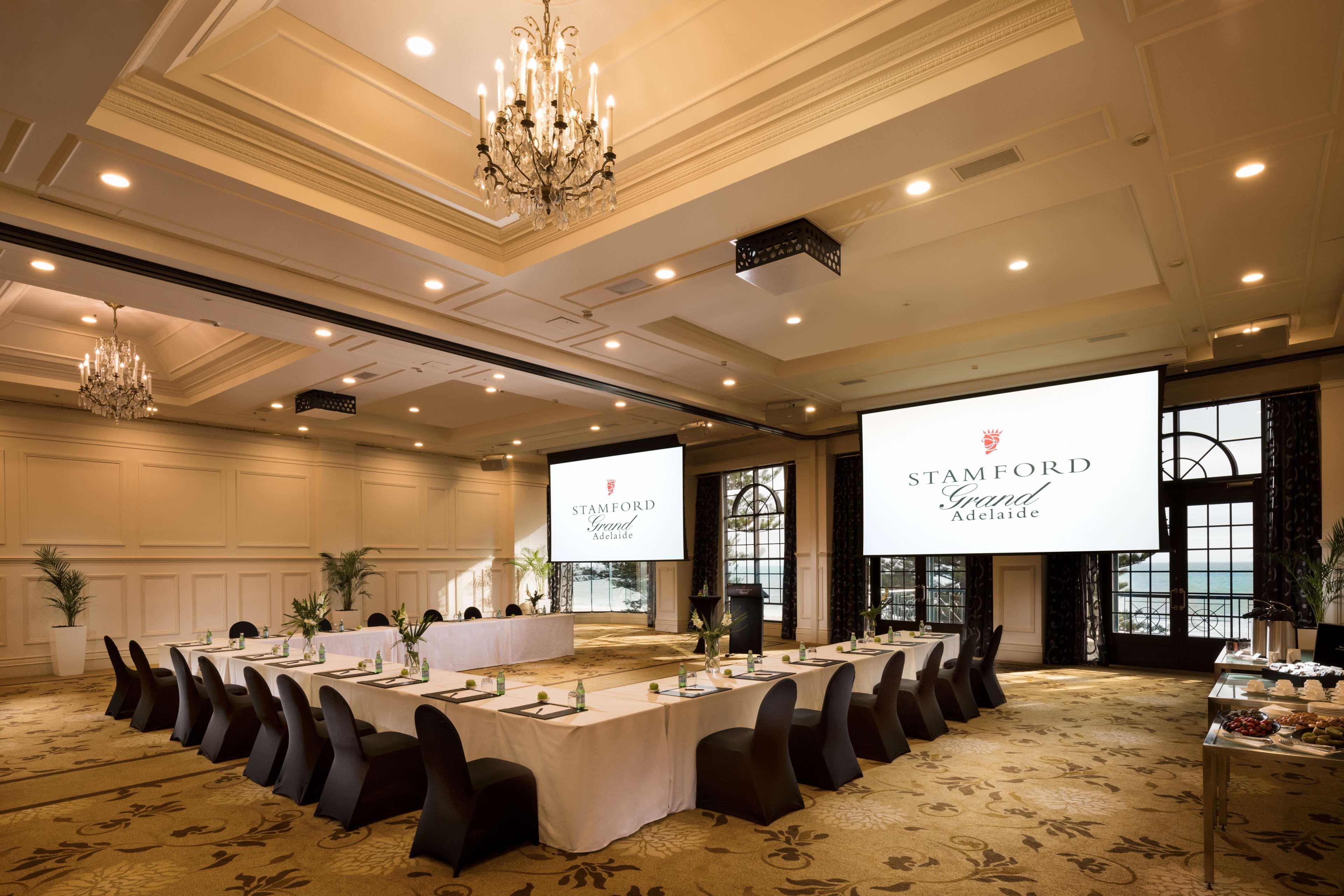 Hotel Stamford Grand Adelaide à Glenelg Extérieur photo The photo shows a spacious and elegantly designed conference room. The room features a long table set for a meeting, surrounded by stylish chairs. Two large screens are positioned at the front, displaying the logo "Stamford Grand Melbourne." The ceil