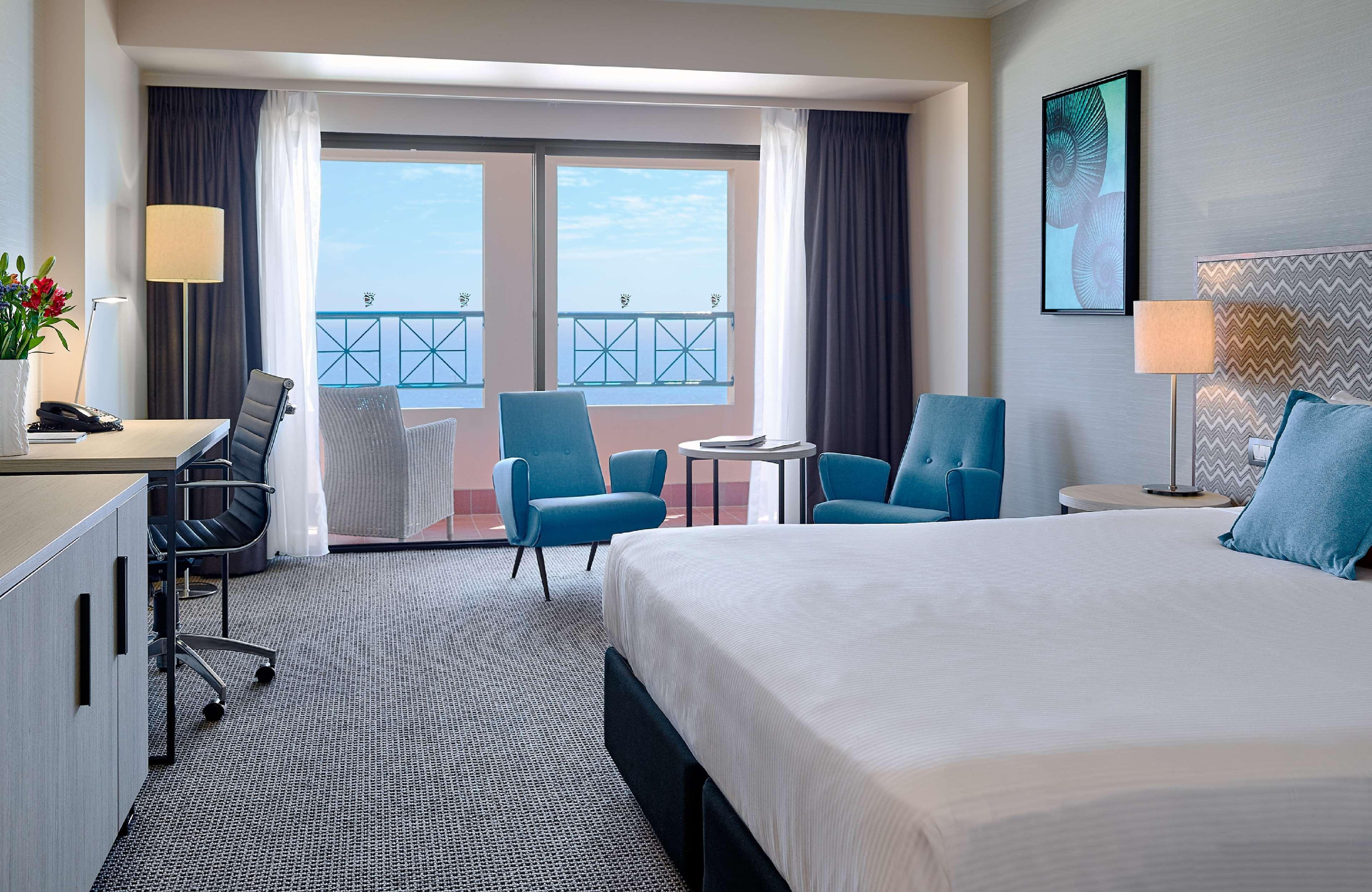 Hotel Stamford Grand Adelaide à Glenelg Extérieur photo The image shows a hotel room featuring a large bed with a white duvet. There are two bright blue chairs and a small table near a large window that provides a view outside, possibly to a balcony. The room includes a workspace with a desk and an office