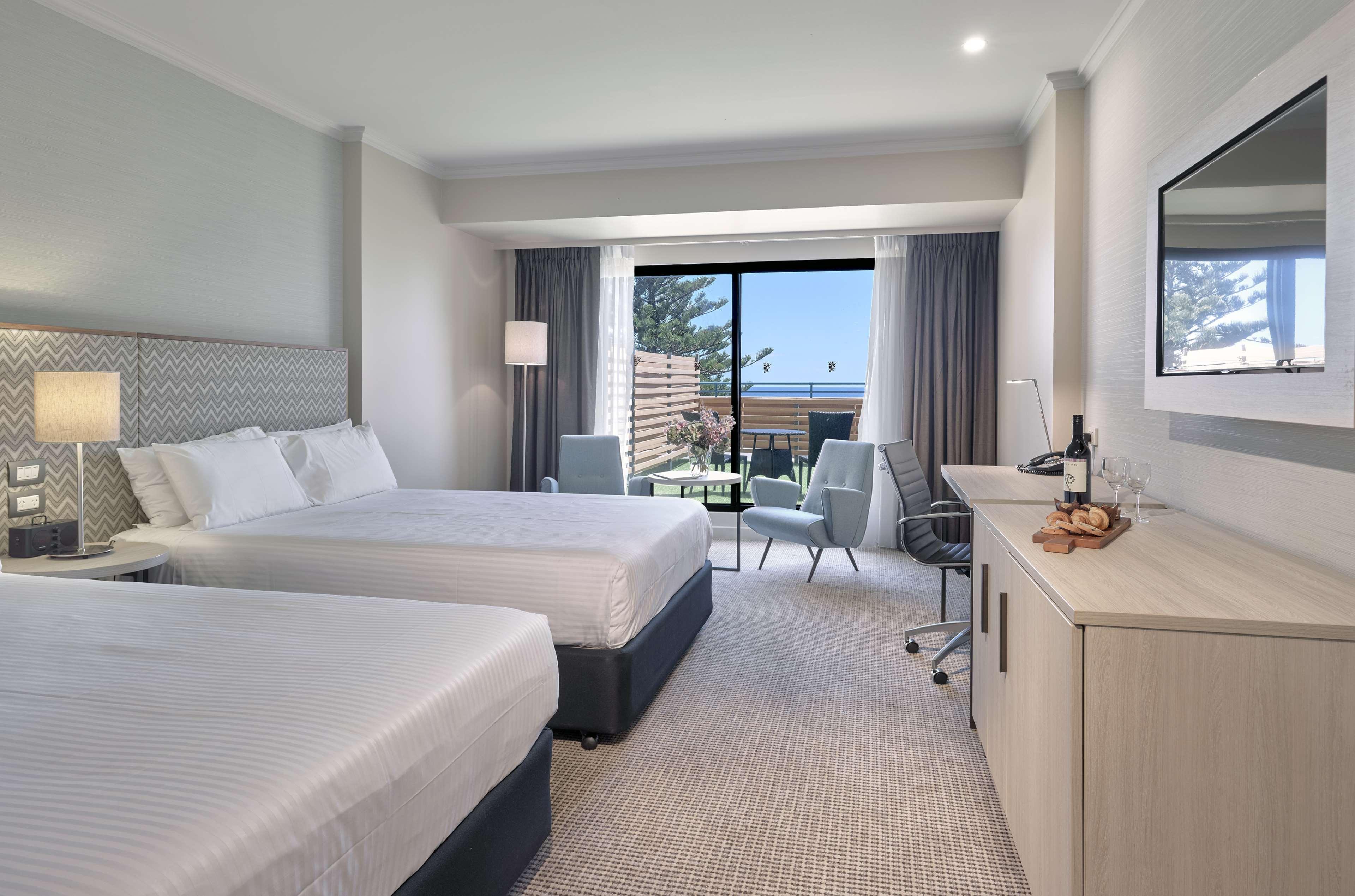 Hotel Stamford Grand Adelaide à Glenelg Extérieur photo The photo shows a modern hotel room featuring two beds with white linens. There's a large window that provides natural light and a view of a balcony area. The room includes two chairs and a small table, creating a cozy sitting area. A television is m