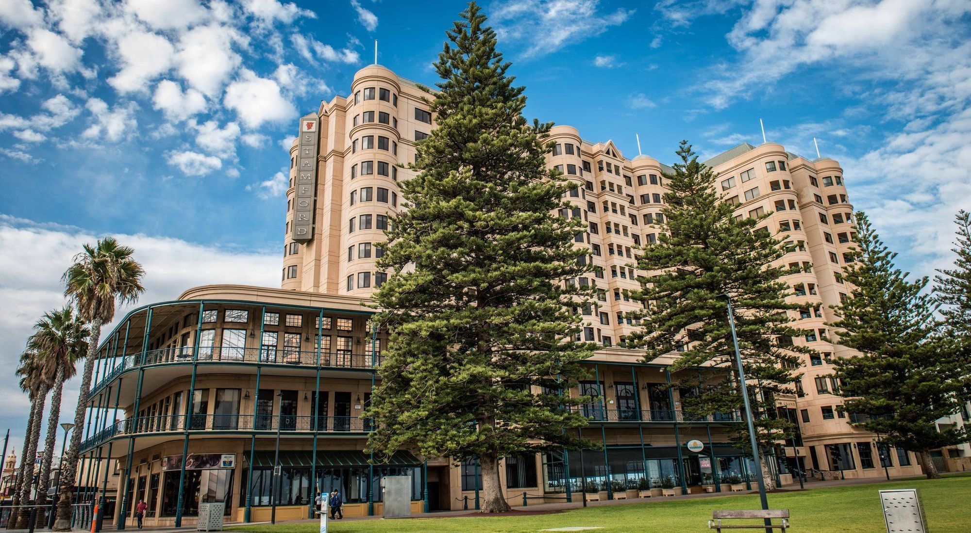 Hotel Stamford Grand Adelaide à Glenelg Extérieur photo The image shows a large building with a distinctive architectural style, characterized by a tan or beige color. The structure appears to have multiple floors and rounded edges. In front of the building, there are several tall green trees, which add t